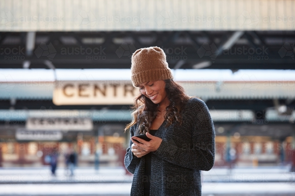 Cheerful Asian woman wearing beanie waiting at train station with mobile phone - Australian Stock Image