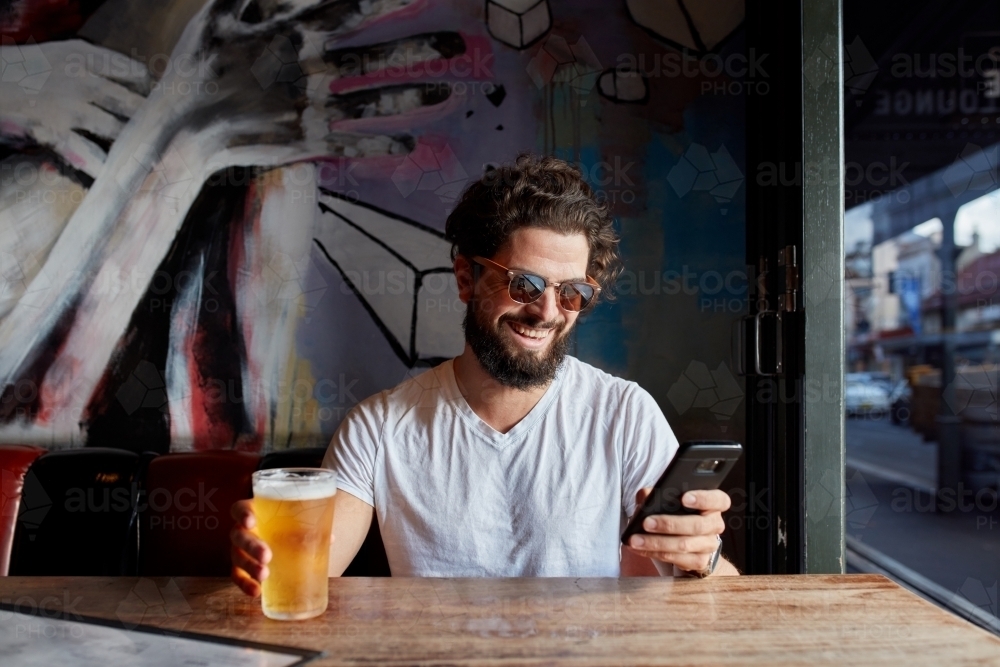 Cheerful hipster man with beer checking mobile phone at bar - Australian Stock Image