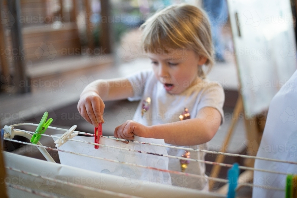 Child carefully pegs a painting to dry at preschool - Australian Stock Image