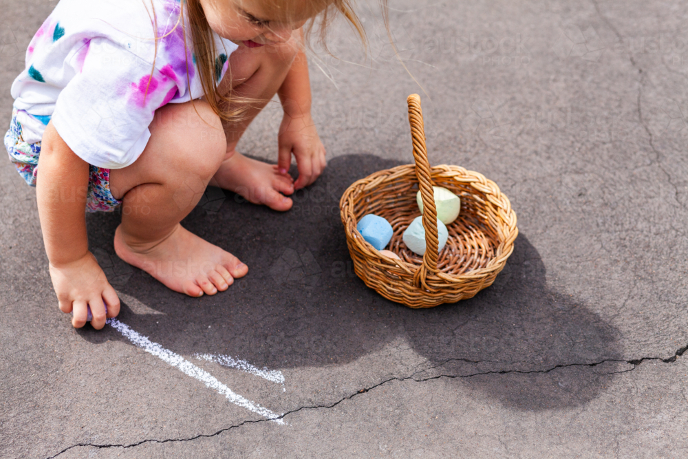 Child crouching down drawing with chalk on driveway outside more chalk in basket - Australian Stock Image