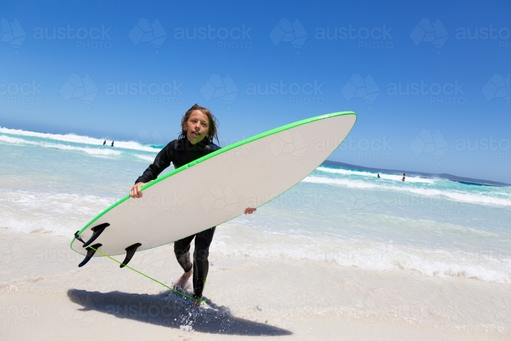 Child in wetsuit carrying surfboard out of the water - Australian Stock Image