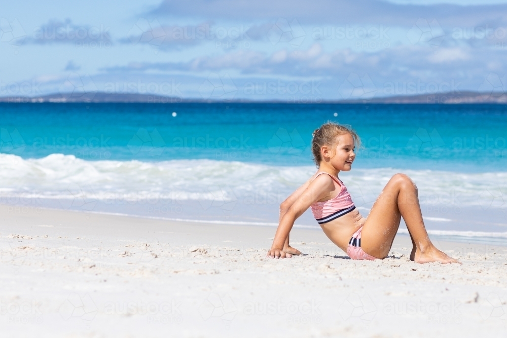 Child sitting on beach with blue water and white sand - Australian Stock Image
