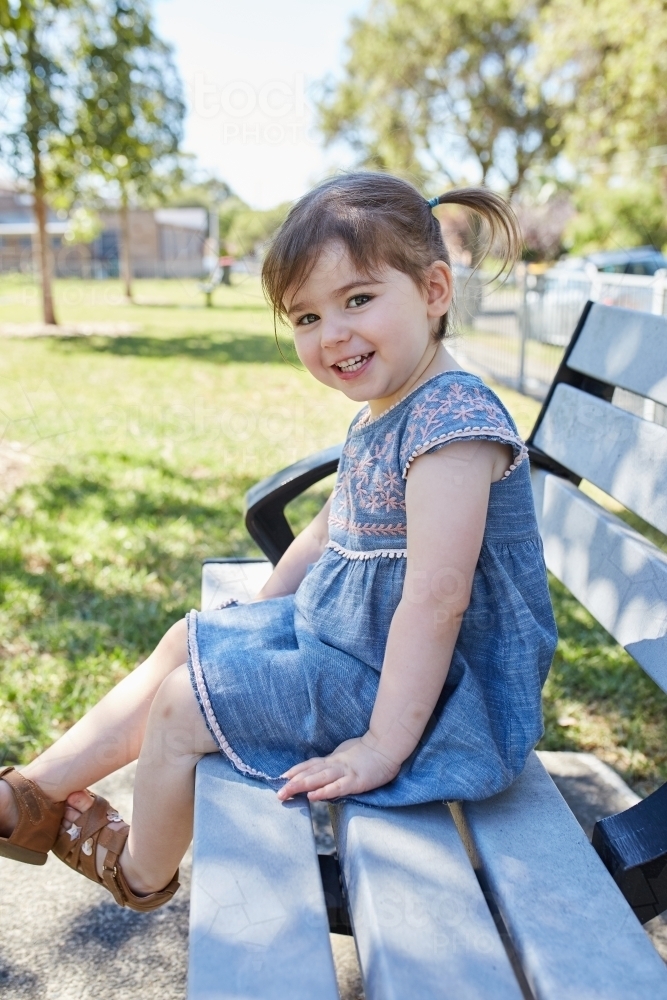 Child smiling on park bench wearing a blue dress - Australian Stock Image