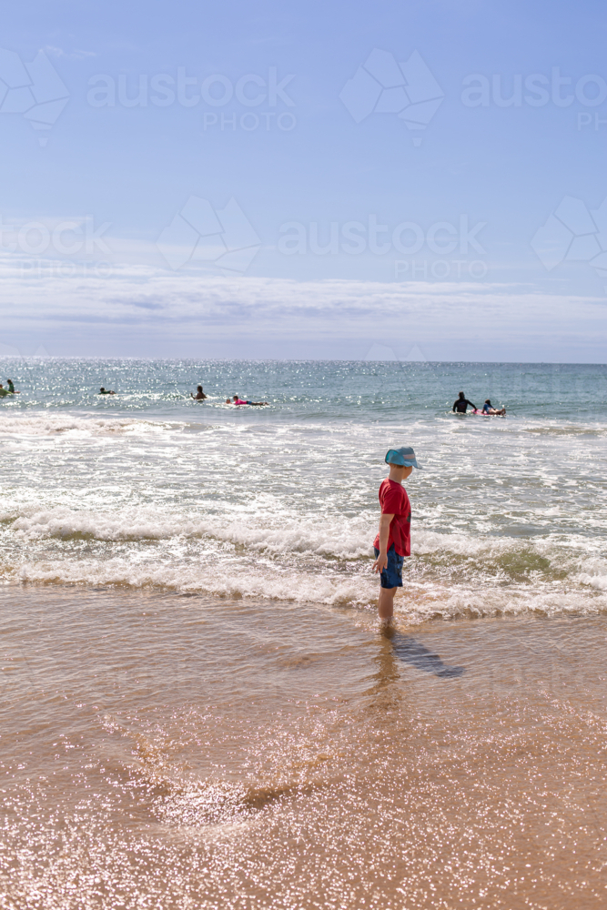 child standing in the shallow water at the beach with surfers in the distance - Australian Stock Image