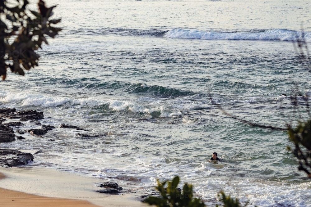 Child swimming in the sea close to shore framed by trees - Australian Stock Image