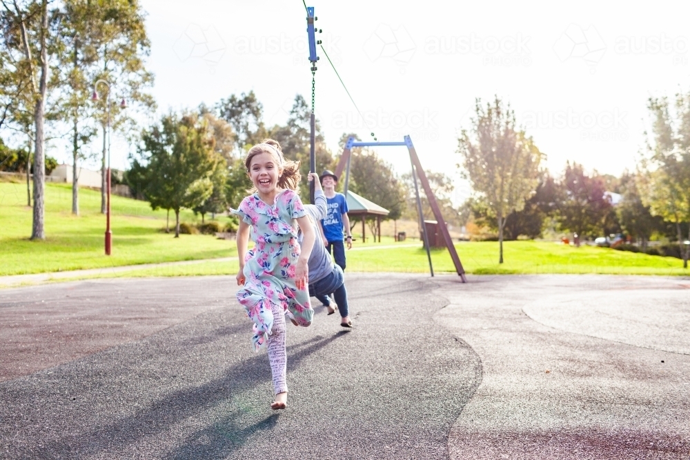 Child, teen and young adult siblings playing together on flying fox swing at playground - Australian Stock Image