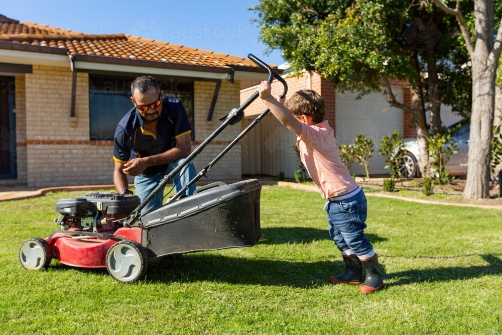 child trying to push lawnmower watched by father - Australian Stock Image