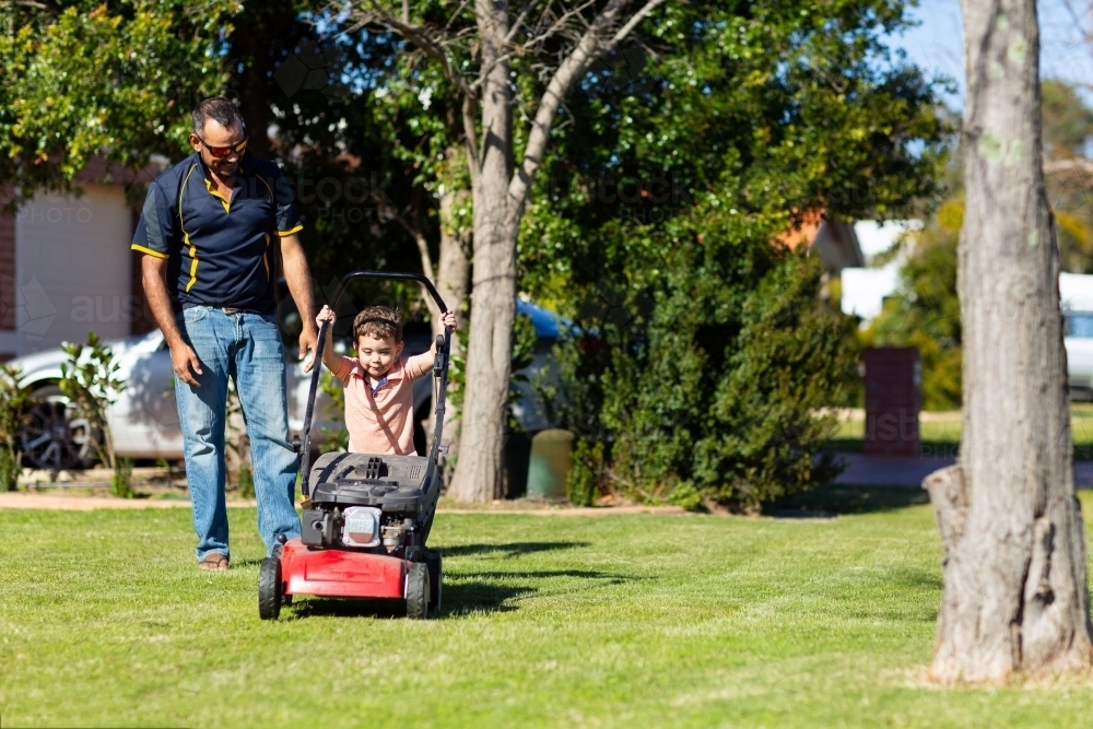 child trying to push lawnmower watched by father - Australian Stock Image