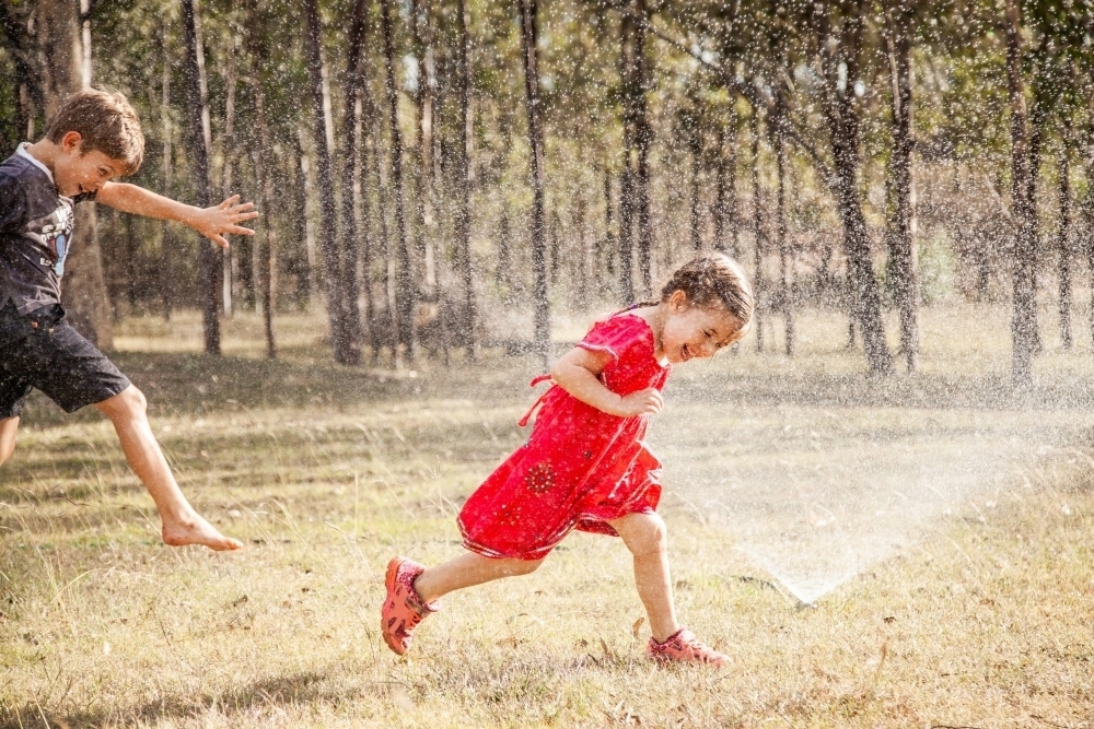 Children running under sprinkler in backyard, Aussie summer childhood - Australian Stock Image