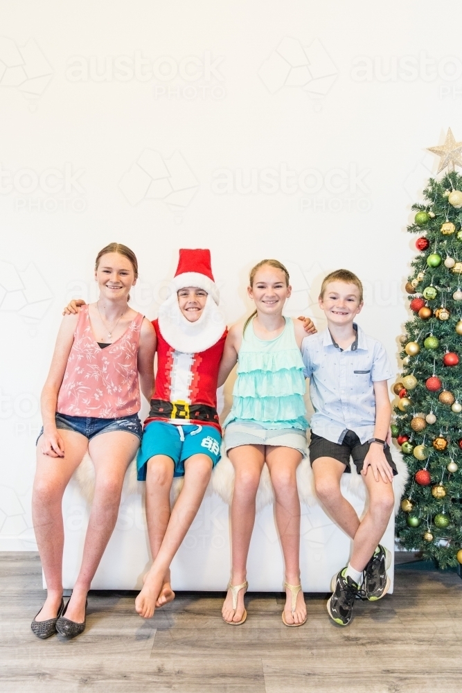 Children sitting together arm in arm next to Christmas tree smiling - Australian Stock Image