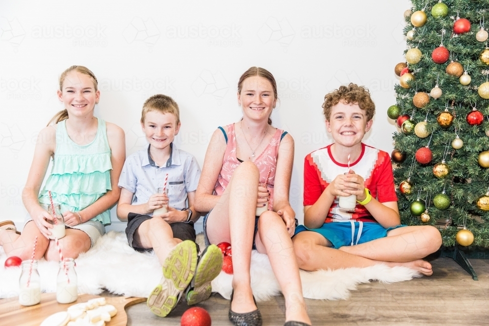 Children sitting together with cookies and milk at Christmas with tree - Australian Stock Image