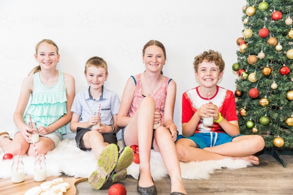 Children sitting together with cookies and milk at Christmas with tree - Australian Stock Image