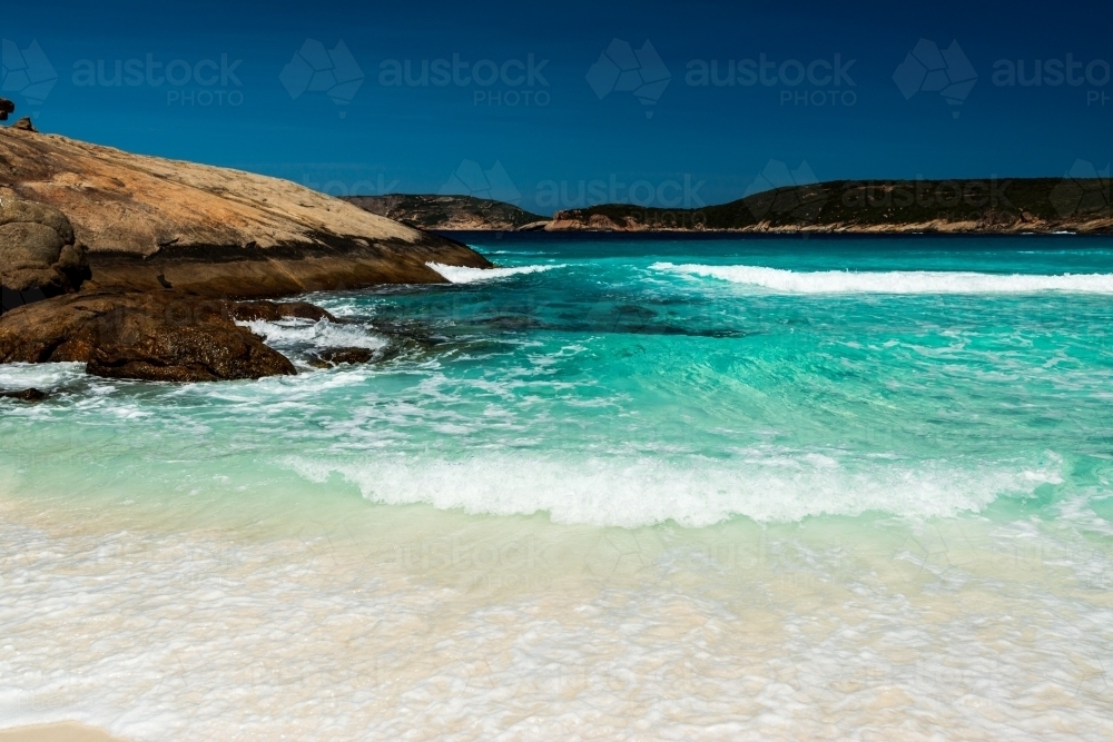 Clear turquoise ocean water washing up on sand with rocky point. - Australian Stock Image