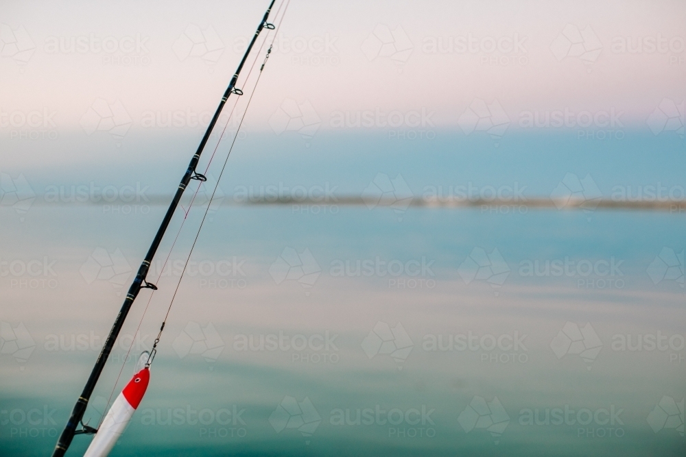 Close-up of a fishing rod with lure with a calm ocean and sky in the background - Australian Stock Image