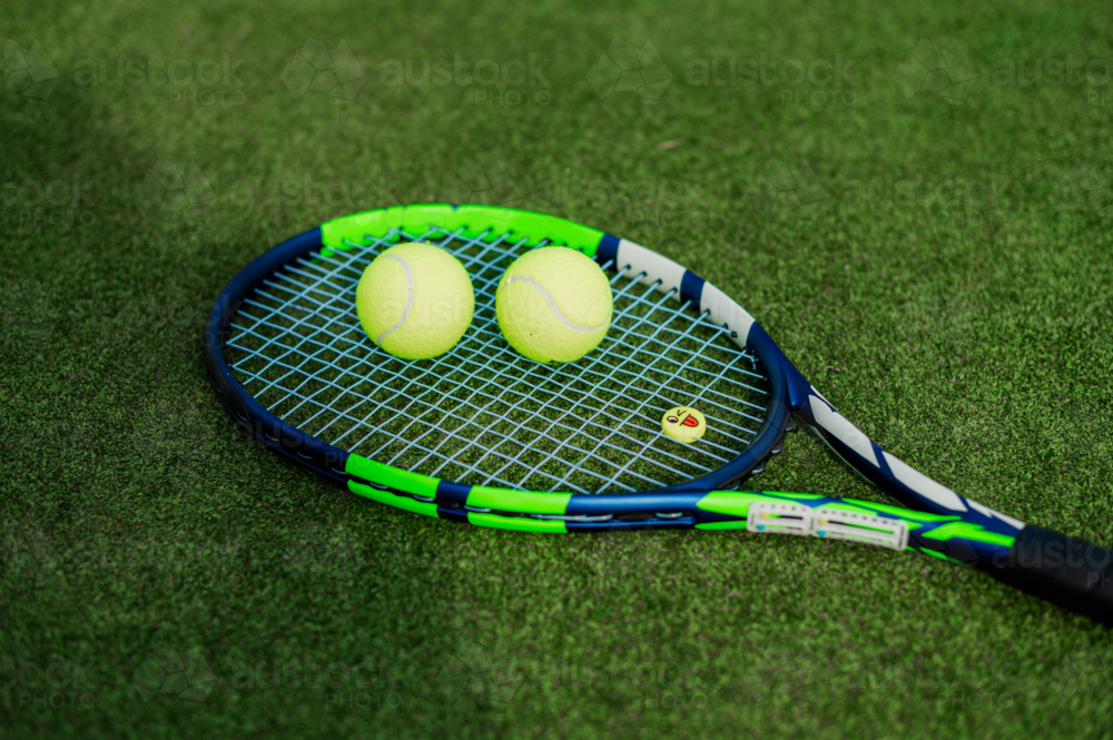 Close-up of a tennis racquet resting on green grass with two yellow tennis balls nearby - Australian Stock Image