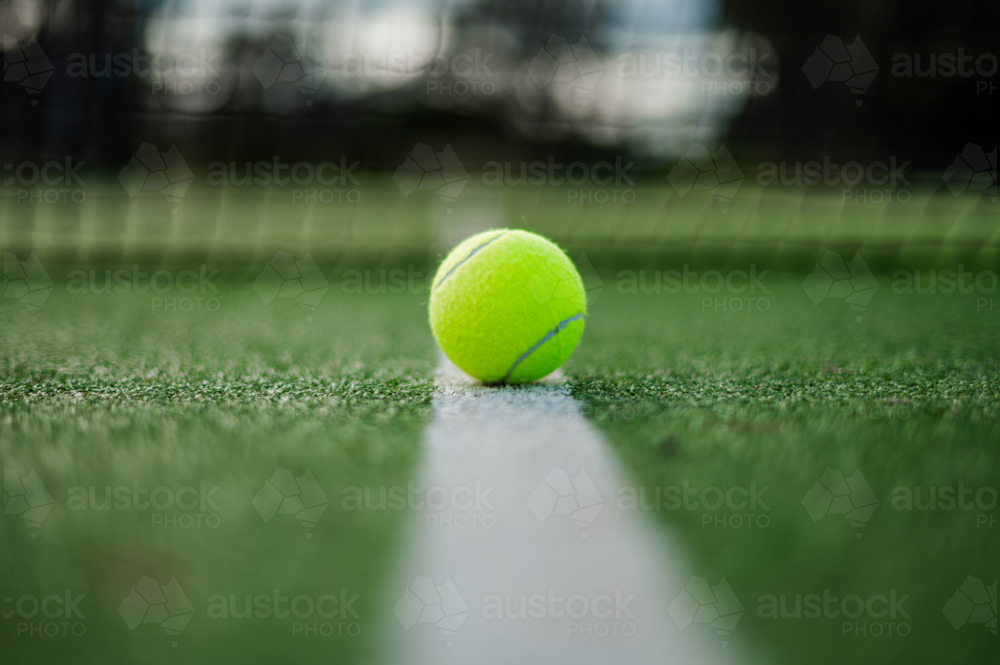 Close-up of a vibrant yellow tennis ball resting on a green court line during daylight - Australian Stock Image