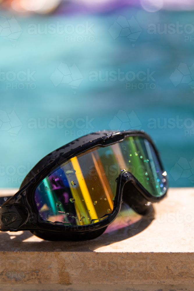 Close-up of black swimming goggles with rainbow-reflective lenses resting on the edge of a pool - Australian Stock Image