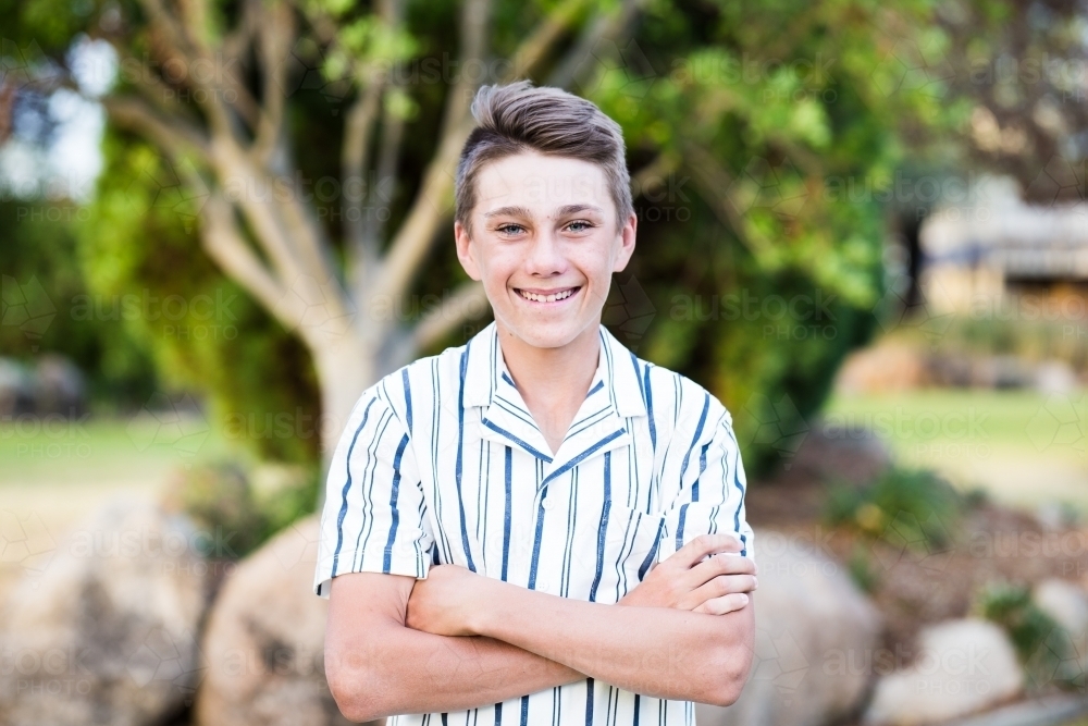 Close up of teenage boy standing in garden smiling with arms crossed - Australian Stock Image