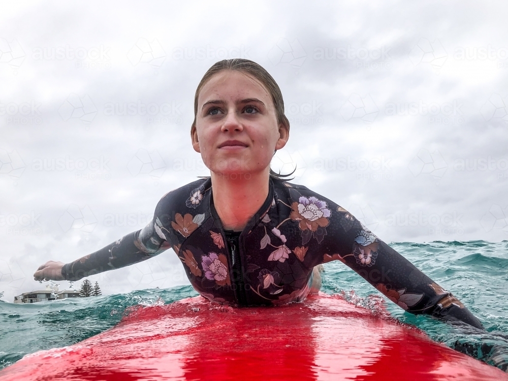 Close up of Teenage girl paddling red surfboard on ocean with beach in background - Australian Stock Image