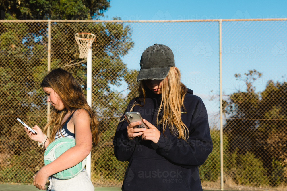 Close up of two teenage girls on smart phones holding ball at outdoor court - Australian Stock Image