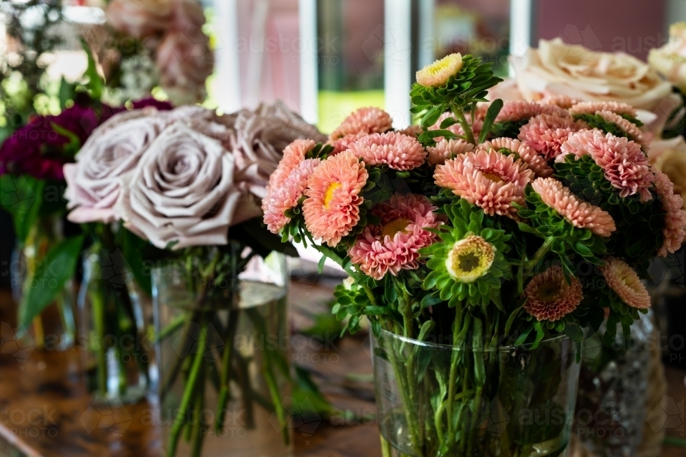 Close up of vase of orange dahlias with blurred vases of mauve and pink roses in the background - Australian Stock Image