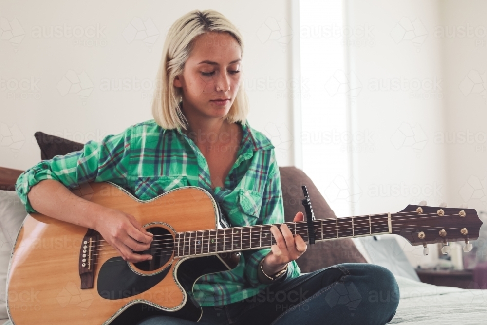 Close up of young blonde woman sitting on bed playing guitar - Australian Stock Image
