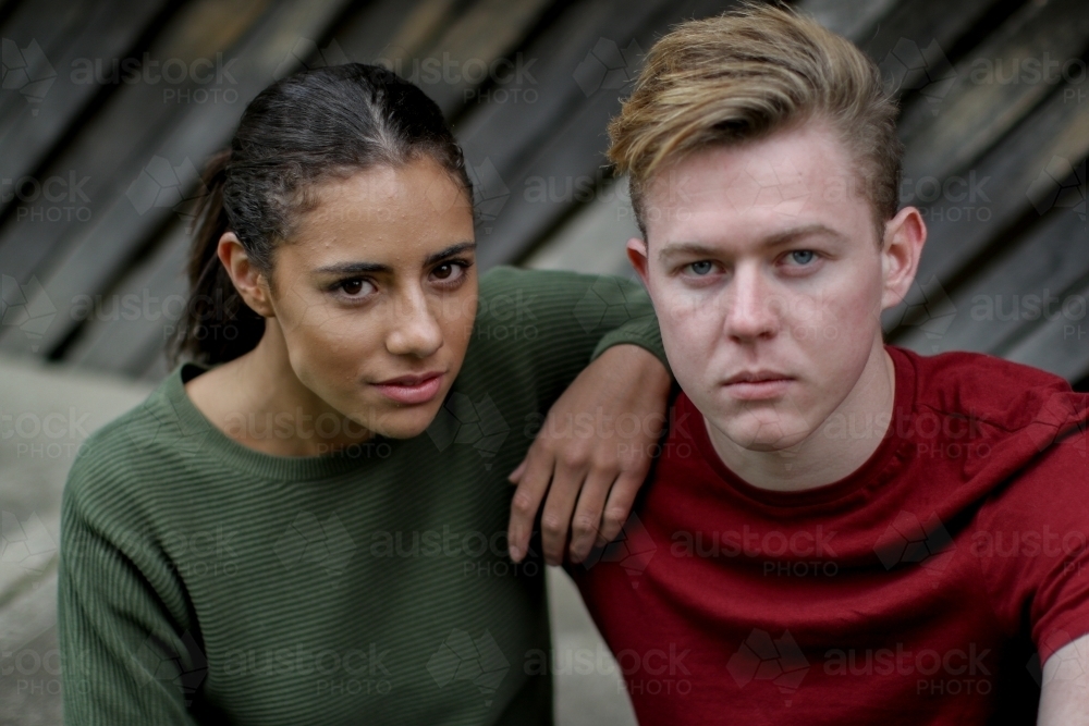 Close up of young interracial couple sitting on ground looking up at camera - Australian Stock Image