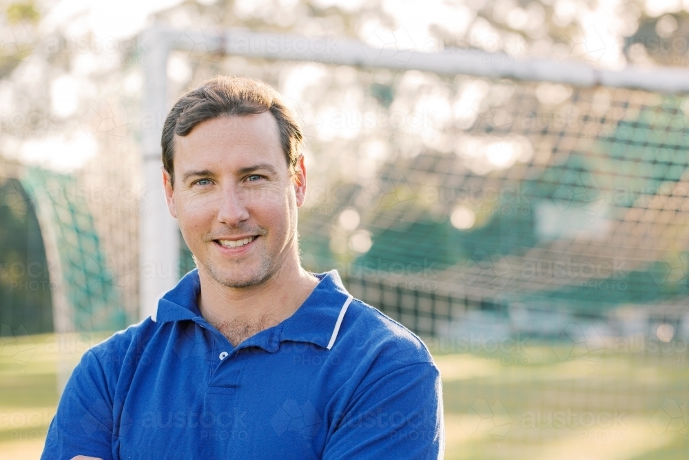 Close up photo of a smiling man wearing blue shirt on a sunny day with goal in the background - Australian Stock Image