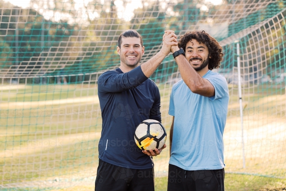 Close up photo of two male friends standing on the field holding and raising their hands with a ball - Australian Stock Image