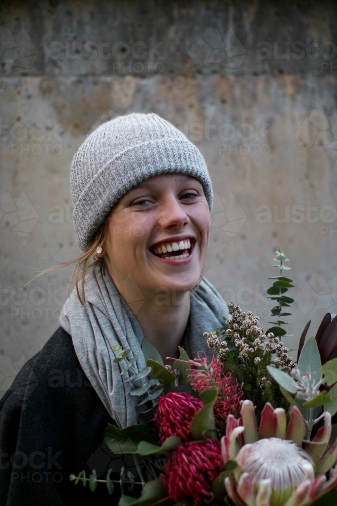 Close up portrait of young girl wearing a beanie and scarf smiling whilst holding a floral bouquet - Australian Stock Image