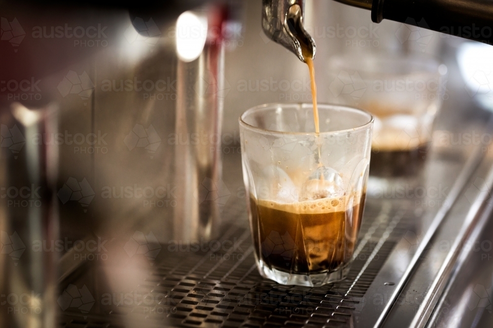 Close up shot of a coffee pouring from a machine into a glass cup on a metal counter - Australian Stock Image