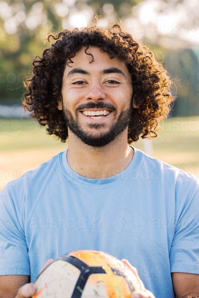 close up shot of a man smiling with curly hair and beard holding a soccer ball wearing blue shirt - Australian Stock Image