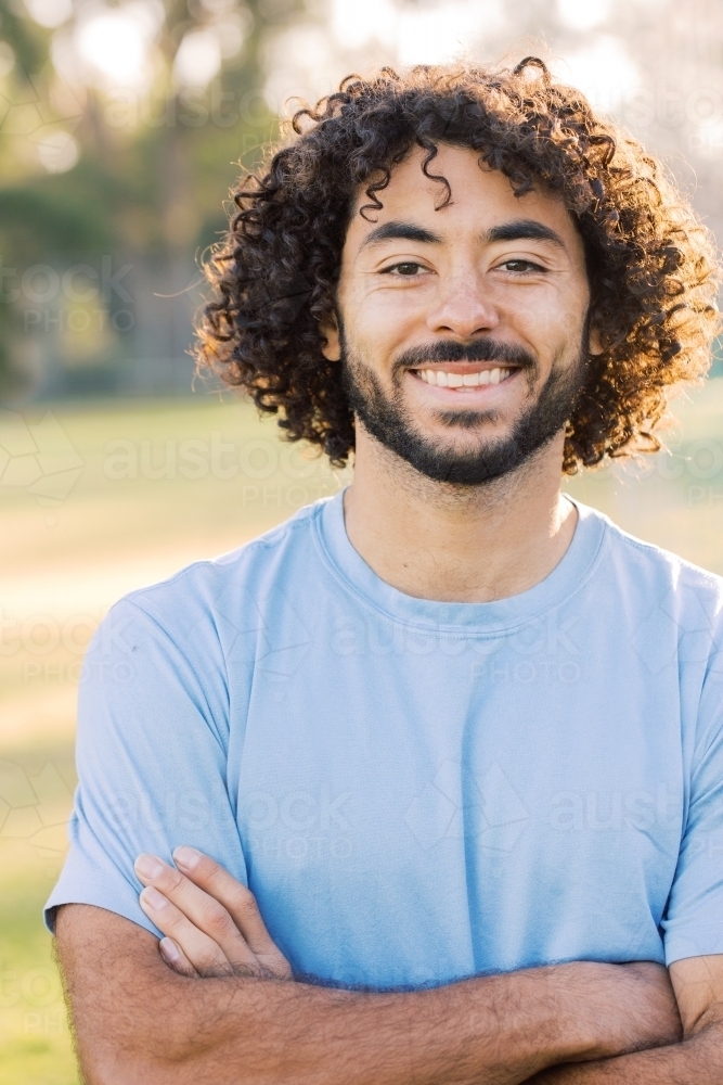 close up shot of a man smiling with curly hair and beard with crossed arms wearing blue shirt - Australian Stock Image