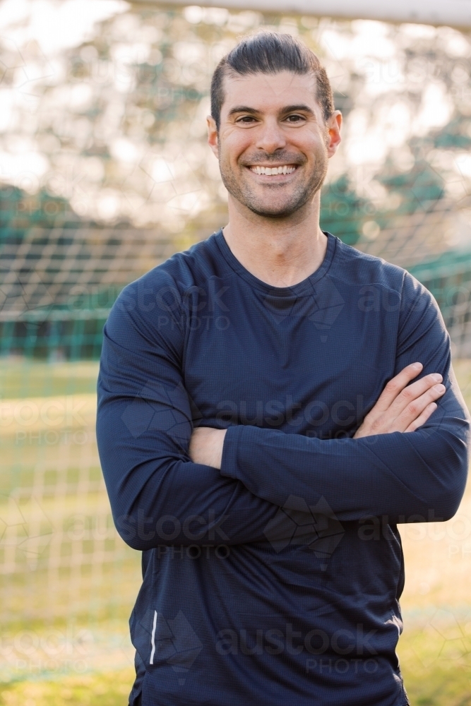 Close up shot of a smiling young man, standing on the  field with crossed hands, wearing blue shirt - Australian Stock Image
