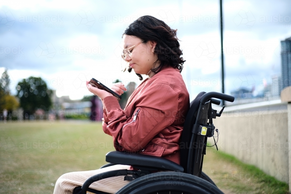 close up shot of a woman with disability sitting in a wheelchair while looking at her mobile phone - Australian Stock Image