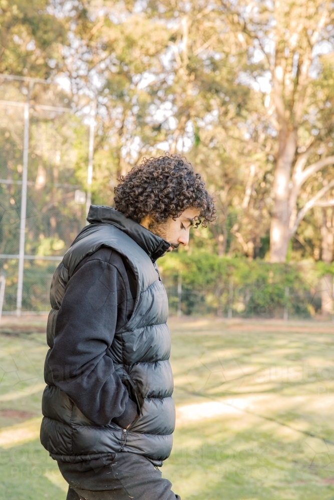 Side view shot of a young men standing on the field with a curly hair looking down with a black vest - Australian Stock Image