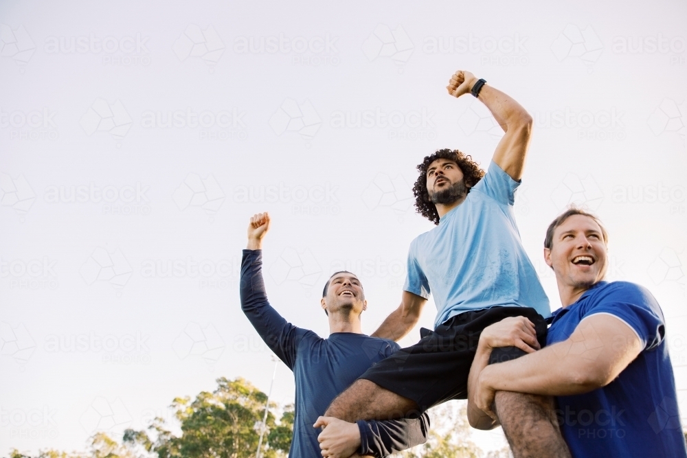 close up shot of two men carrying a man on their shoulders while raising their arms up high - Australian Stock Image