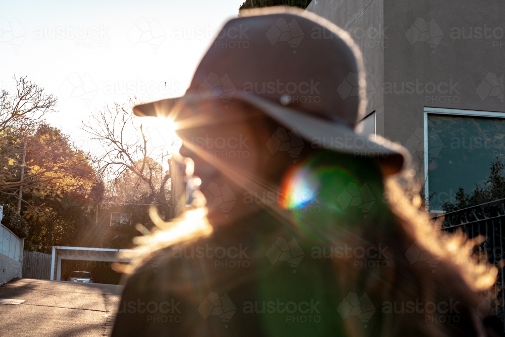 close up shot of woman with eyes closed wearing a hat and a house and sunlight in the background - Australian Stock Image