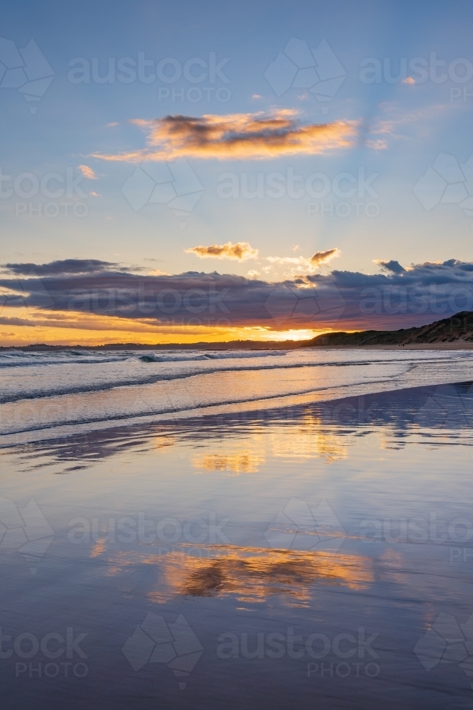 Cloud formation and sunset reflected on a wide sandy beach with gentle waves - Australian Stock Image
