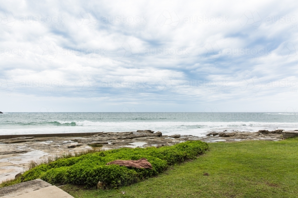 Cloudy skies over beach with ocean waves behind grass - Australian Stock Image