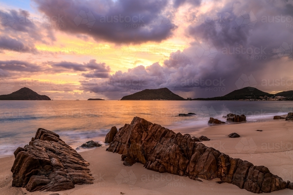 Cloudy sunrise over mountains and rocky beach - Australian Stock Image