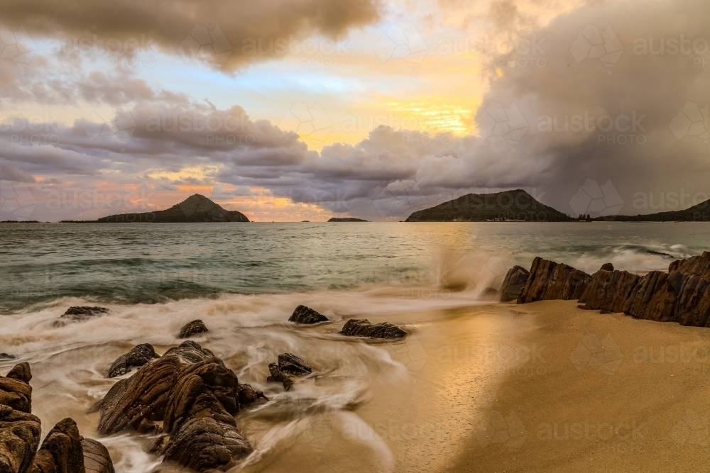 Cloudy sunrise over mountains and rocky beach - Australian Stock Image