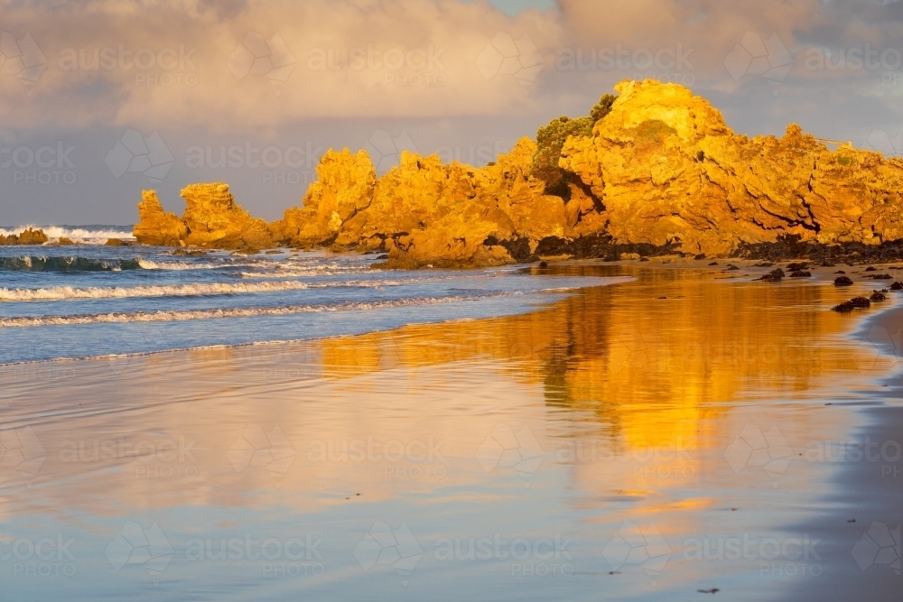 Coastal rock formations in golden lighting reflected on a wet sandy beach - Australian Stock Image