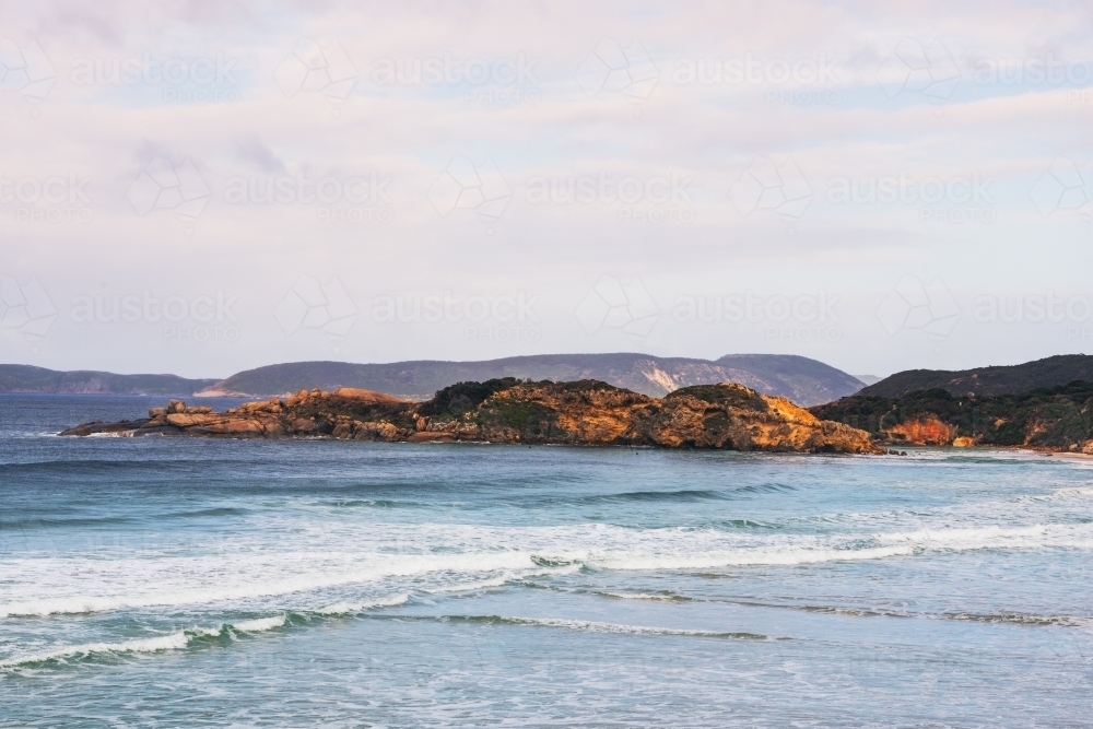 Coastal shot of beach with rocky headland and two surfers sitting in water - Australian Stock Image