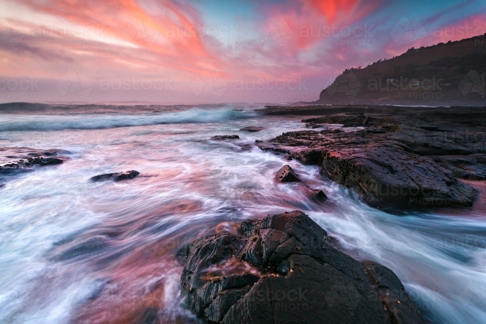 Colossal waves wash onto the coast, with sunrise colouring the clouds - Australian Stock Image