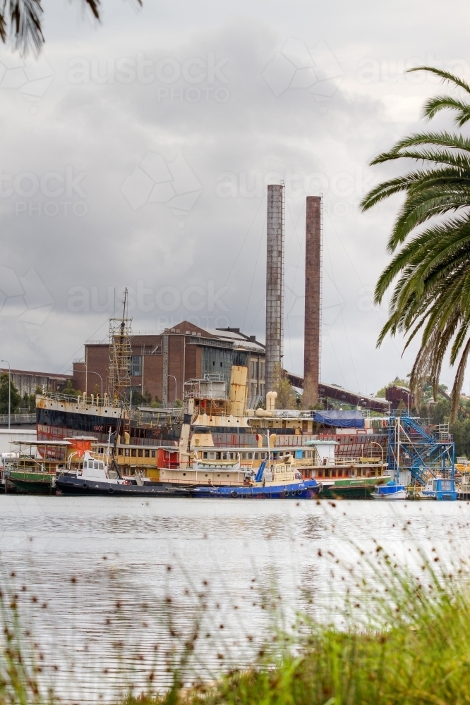 Colourful derelict boats across the water with run down industrial building in the background - Australian Stock Image