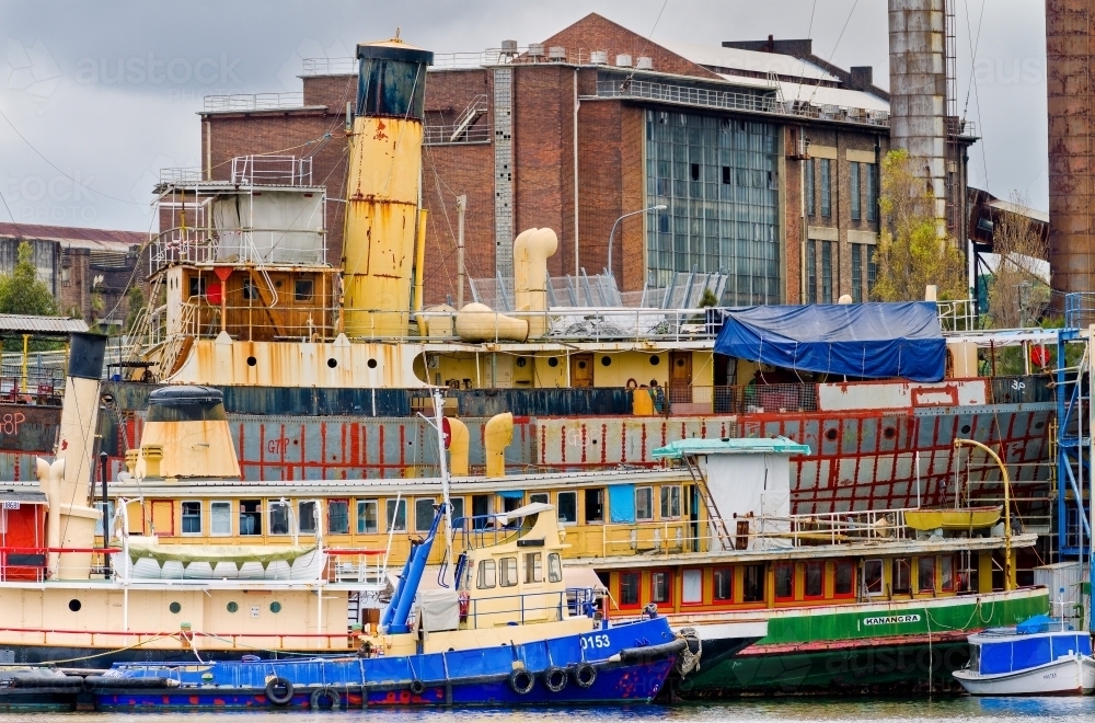 Colourful derelict boats and run down industrial building in background with smashed windows - Australian Stock Image