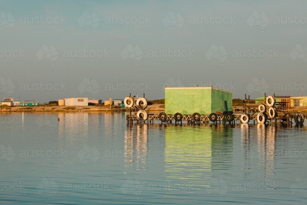 Colourful fishing shacks and shed with wooden jetty and old tyres, reflected in calm water - Australian Stock Image