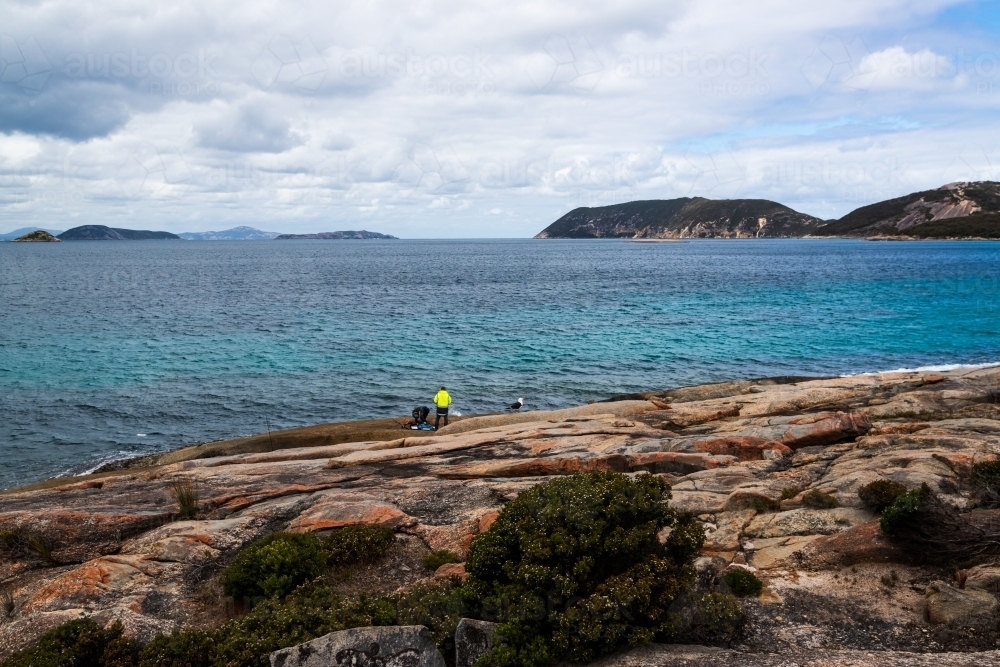 Colourful rocky foreshore with fishermen and dark blue sea - Australian Stock Image