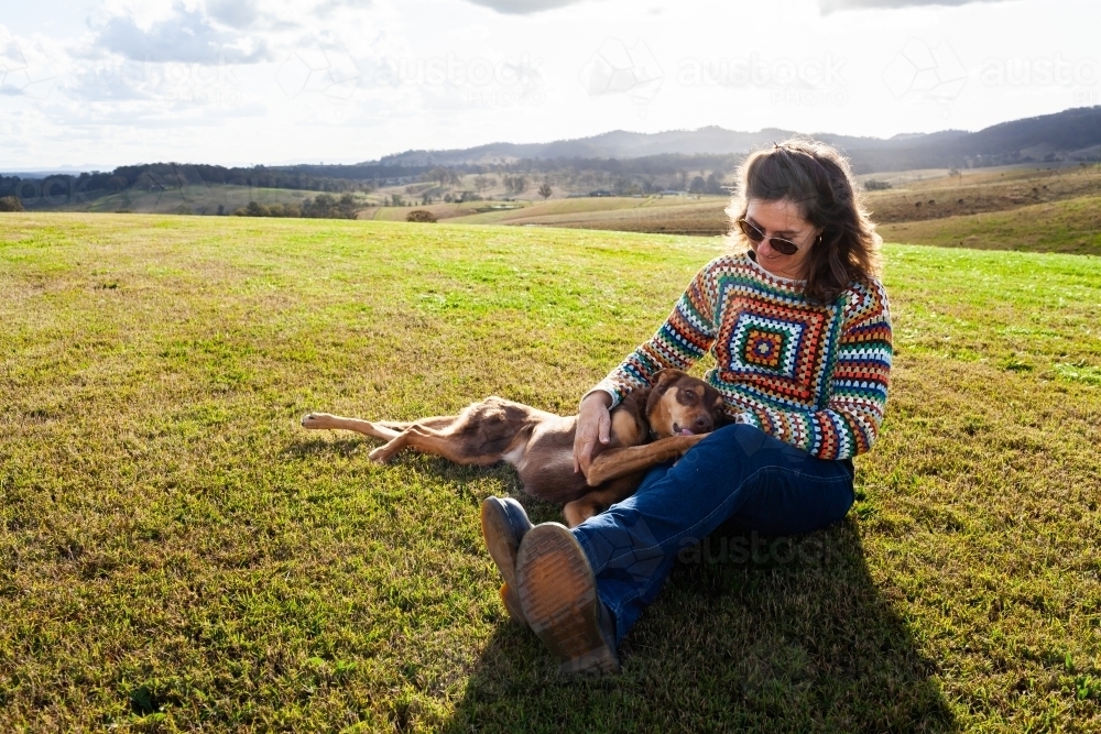 Companions - happy middle age woman and kelpie dog sitting together on farm hilltop - Australian Stock Image